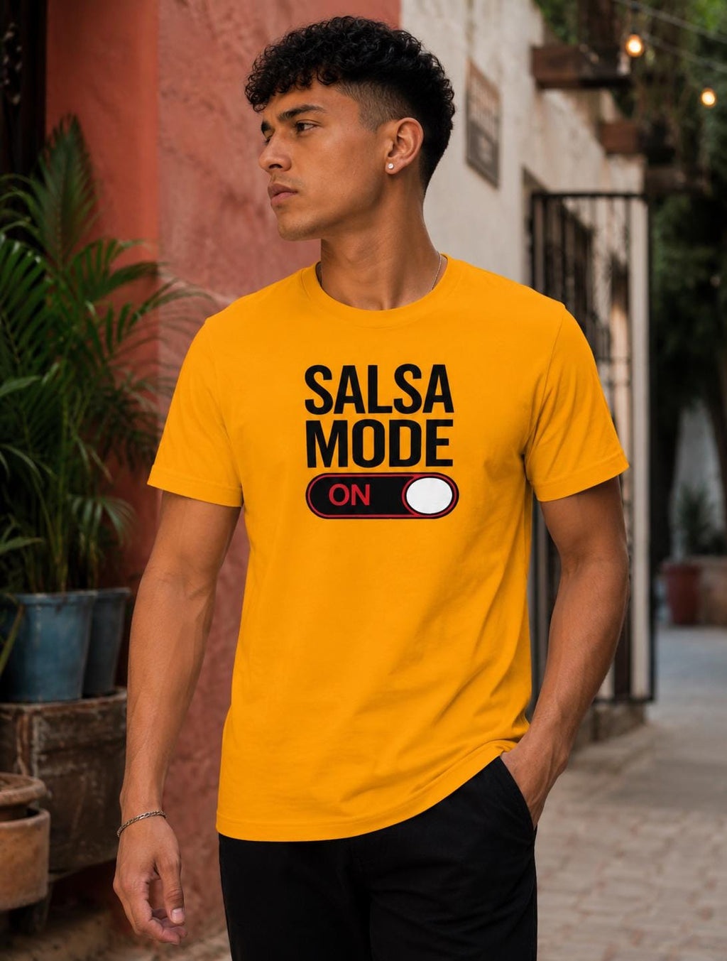 Male model wearing a bright yellow “Salsa Mode ON” graphic t-shirt with bold black lettering and switch design, styled casually in an outdoor street setting. Fun salsa dance apparel for Latin dance lovers, social dancers, and salsa lifestyle fashion.